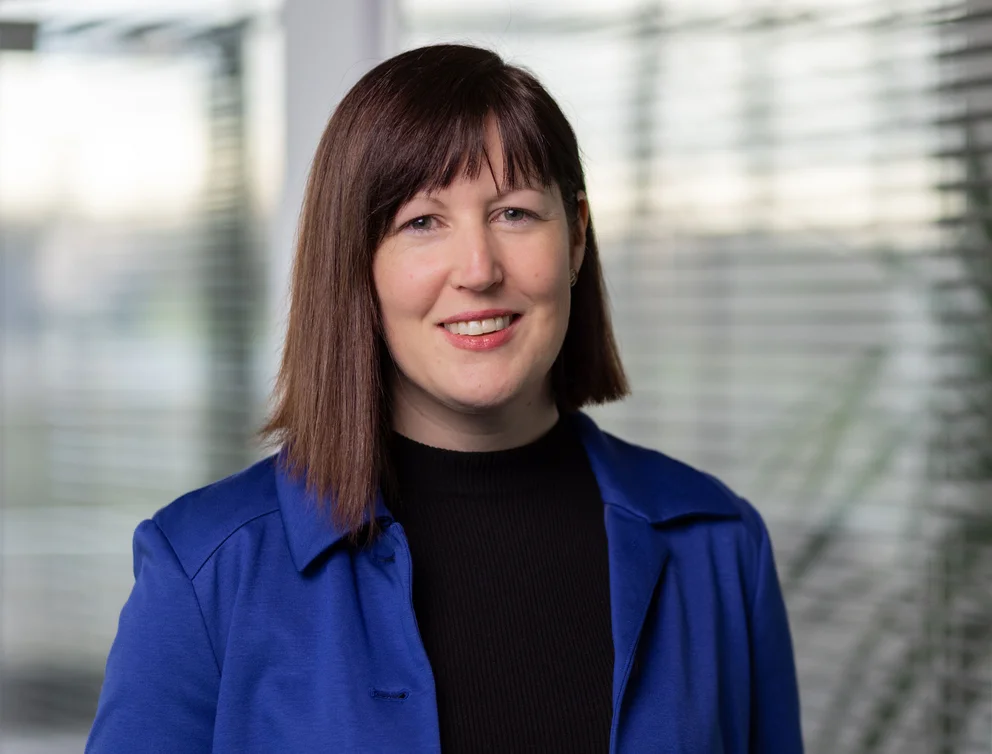 A professional woman in a blue blazer stands in an office, representing PVA TePla expertise in semiconductor equipment and vacuum technology.